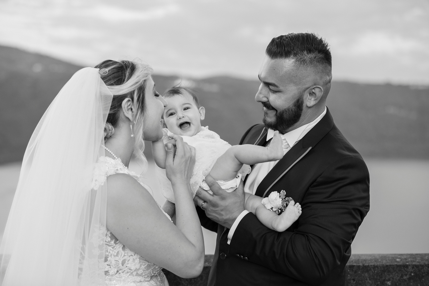 In riva al Lago di Castel Gandolfo, una foto sposo, sposa e il loro bimbo. Il fotografo di matrimonio, cattura l'amore familiare, arricchendo il servizio fotografico con la bellezza del luogo. Uno scatto commovente durante il servizio fotografico